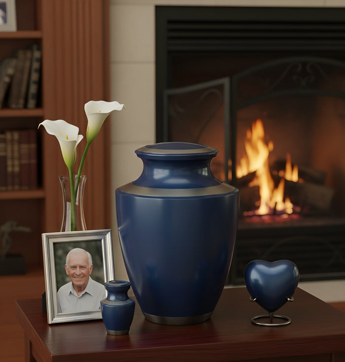 Deep Blue Serenity Collection displayed on a dark wood mantle beside a lit fireplace — navy blue adult cremation urn, mini keepsake urn, and heart keepsake with framed portrait and white calla lilies