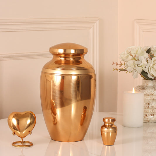 Polished bronze Art Deco cremation urn set with large adult urn, small keepsake heart urn on stand, and mini token urn, displayed with white flowers and candle